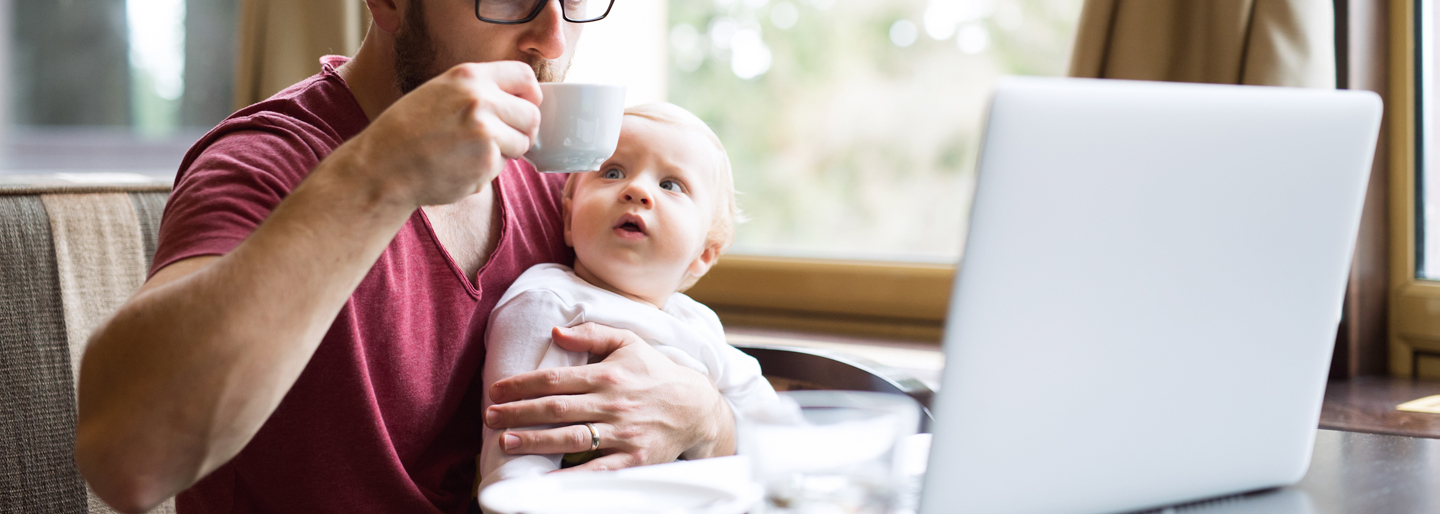 A dad having coffee at his a computer with his baby.